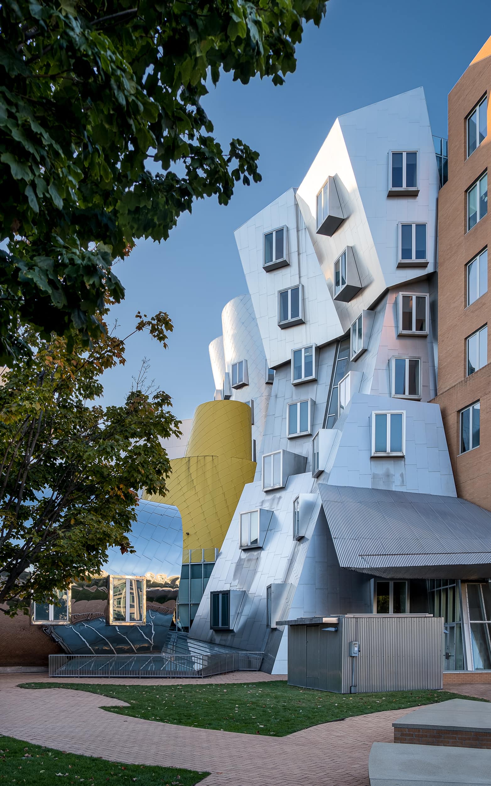 Architectural photography of the Ray and Maria Stata Center at the Massachusetts Institute of Technology (MIT) in Boston designed by Frank Gehry featuring a side angle capturing the rear of the building in brick and aluminum with a pop of yellow framed by a large tree on the left side of the frame
