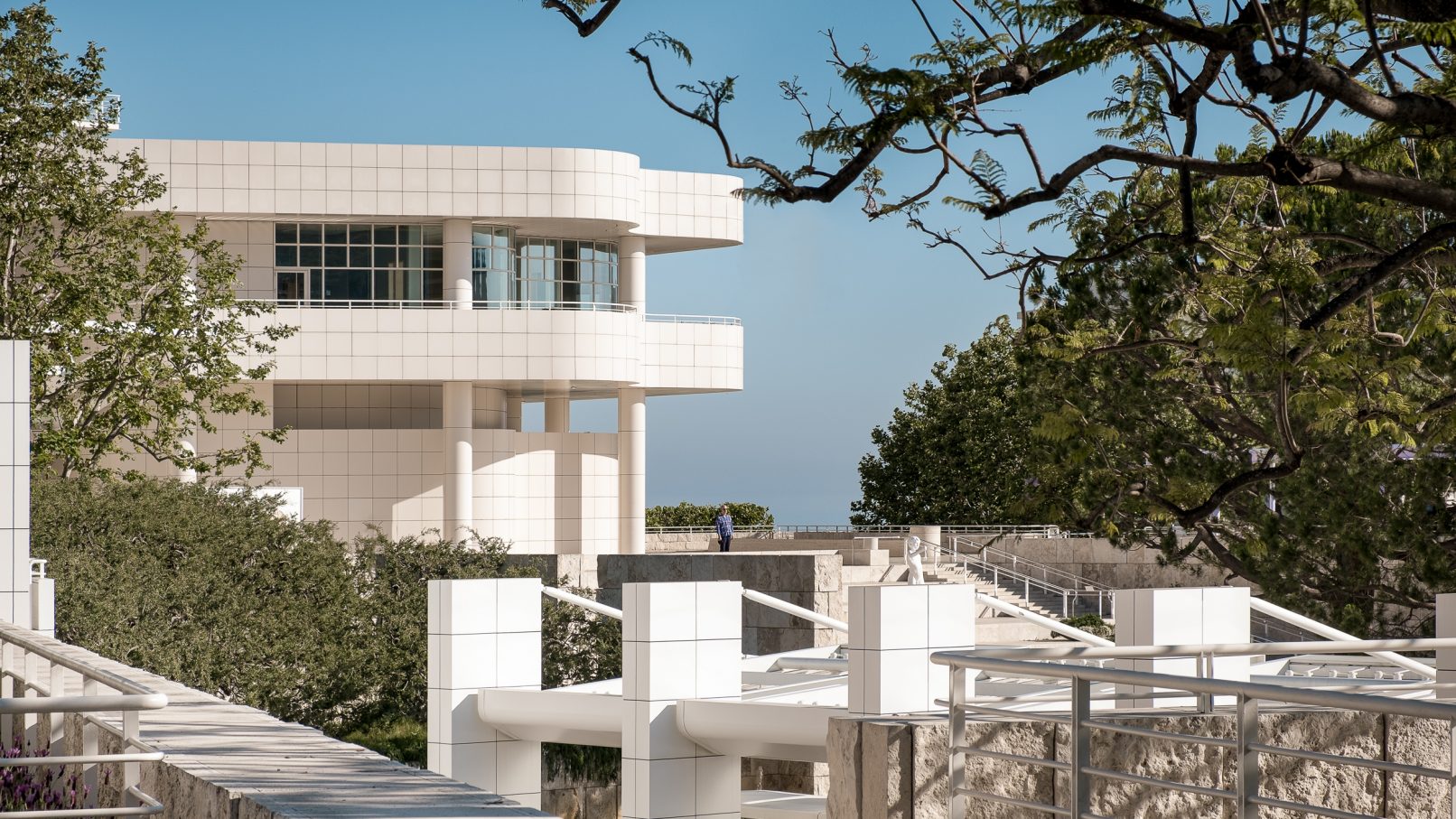 Architectural Photograph of the Getty Center in Los Angeles designed by Richard Meier and Partners featuring a long a shot back towards the main Getty Center building just above where the tram departs framed by the long branches of a tree with only a single onlooker in the shot