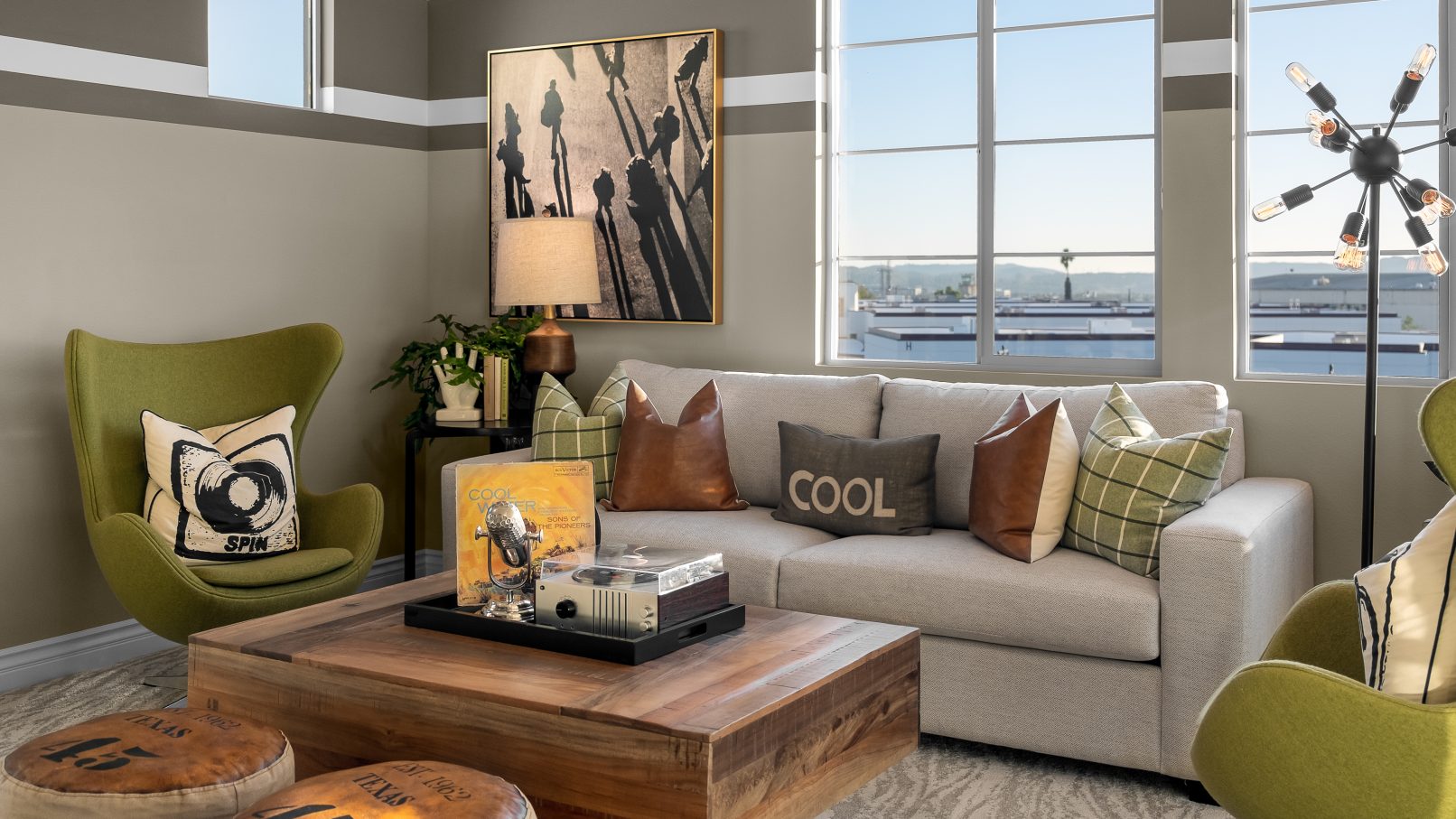 Orange County Interior Design Photography of a Living Room with a Grey Couch and Green Accent Chairs and a Wood and Leather Coffee Table and Large Panoramic Windows with Dramatic Wall Art and a Pillow on the Couch that says Cool